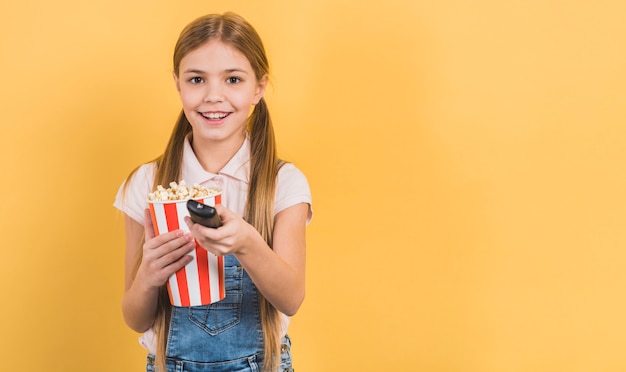 Smiling girl holding popcorn in hand changing the channel with remote control against yellow background