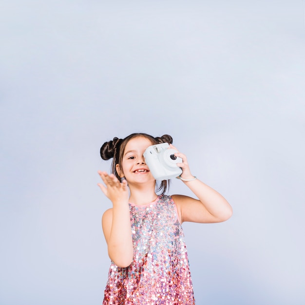 Smiling girl holding retro instant camera in front of her one eyes against blue backdrop