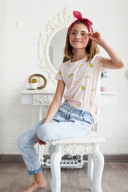 Smiling girl holding spectacle and looking at camera while sitting on white wooden table