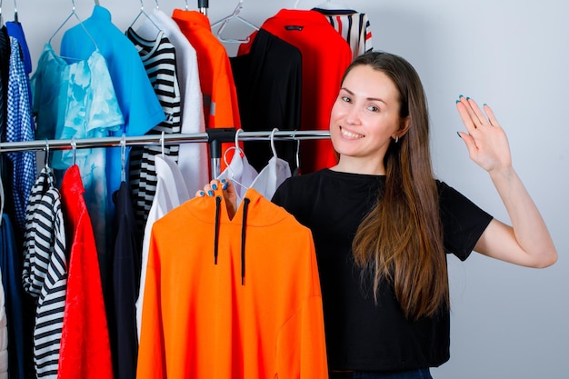 Smiling girl is holding sweetshirt and showing hi hesture with other hand on clothes background