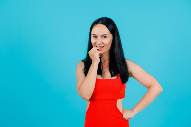 Smiling girl is holdinh one hand on lips and other on waist on blue background