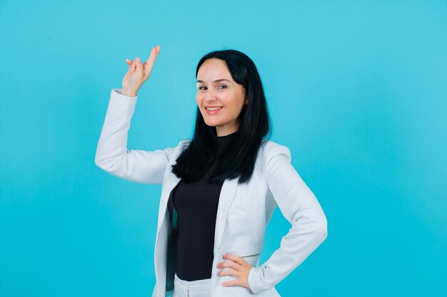 Smiling girl is looking at camera by crossing fingers and putting hand on waist on blue background