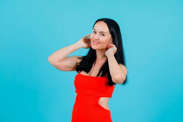 Smiling girl is looking at camera by holding hands near ears on blue background