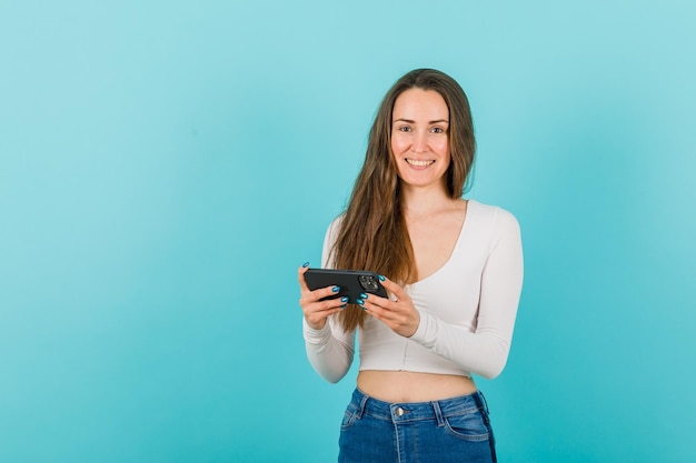 La ragazza sorridente sta guardando la fotocamera tenendo lo smartphone su sfondo blu