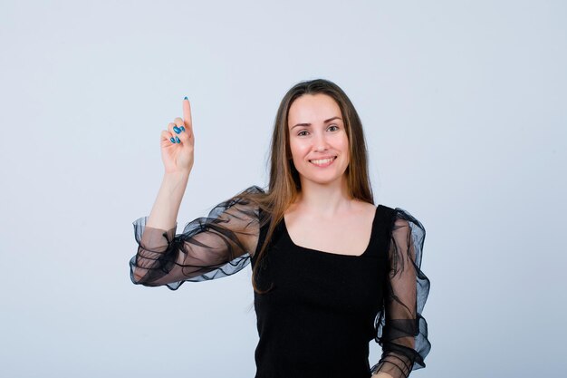 Smiling girl is looking at camera by pointing up with forefinger on white background