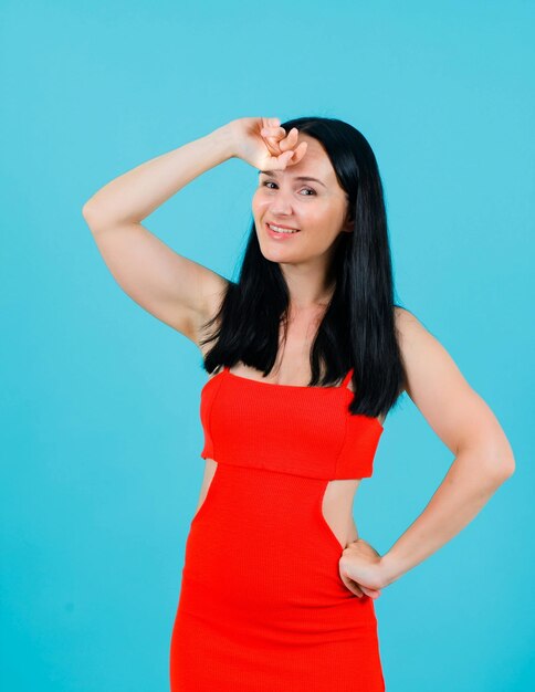 Smiling girl is looking at camera by putting one hand on forehead and other on waist on blue background