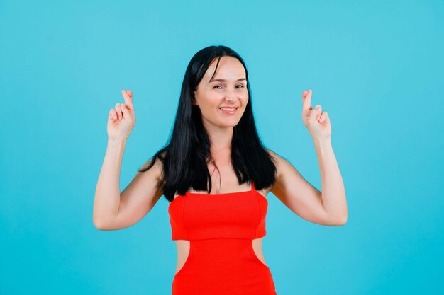 Smiling girl is looking at camera by raising up her crossed fingers on blue background