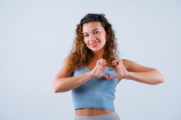 La ragazza sorridente sta guardando la fotocamera mostrando il gesto del cuore su sfondo bianco