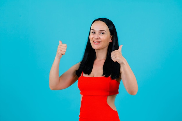 Smiling girl is looking at camera by showing perfect gestures with thumbs on blue background