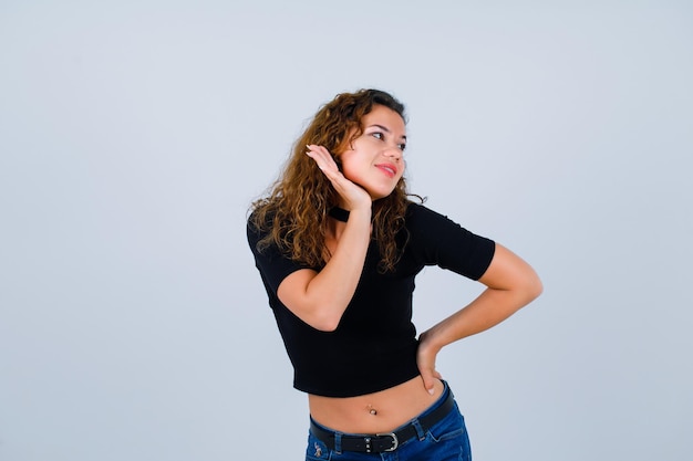 Smiling girl is looking left by holding hand under chin and putting other hand on waist on white background