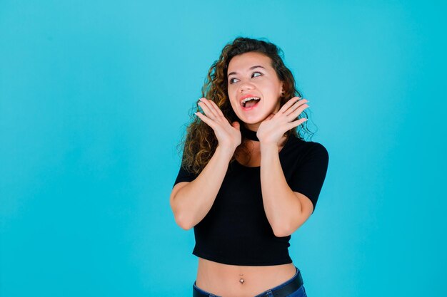 Smiling girl is looking left by holding hands under chin on blue background