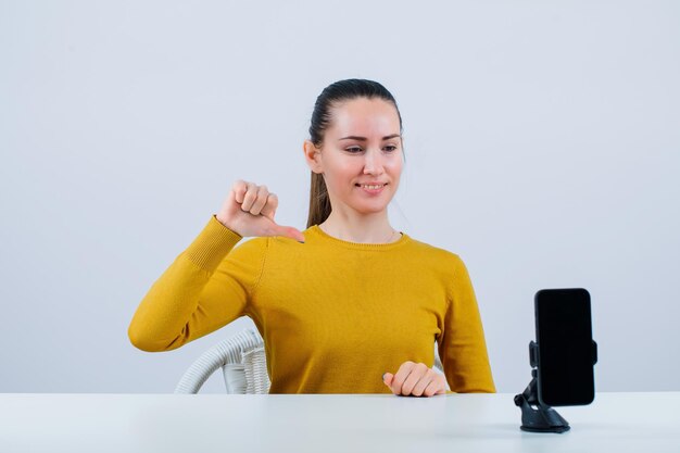 Smiling girl is pointing down with thumb by sitting in front of mobile camera on white background