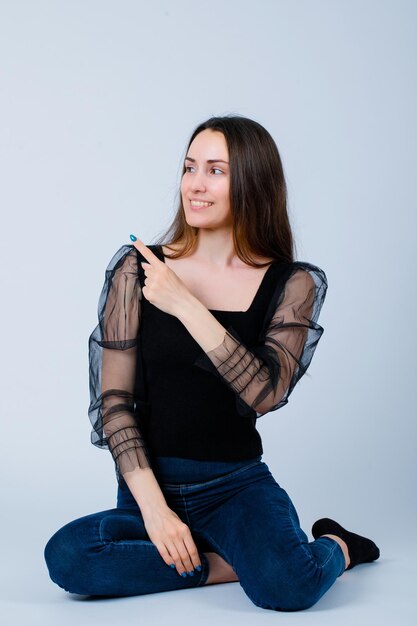 Smiling girl is pointing left with forefinger by sitting on floor on white background