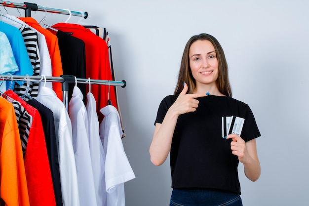Smiling girl is pointing right and holding credit cards on clothes background