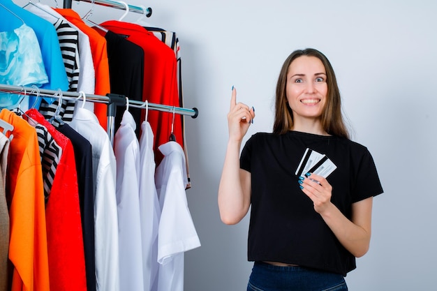 Smiling girl is pointing up with forefinger and holding credit cards on clothes background