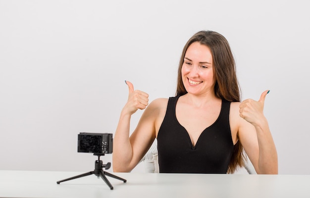 Улыбающаяся девушка позирует перед маленькой камерой, показывая идеальные жесты на белом фоне