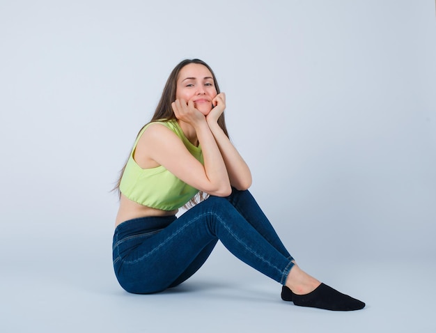 Smiling girl is putting hands under chin by sitting on floor on white background