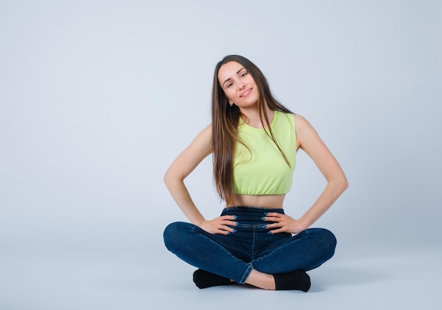 Smiling girl is putting hands on waist by sitting on floor on white background