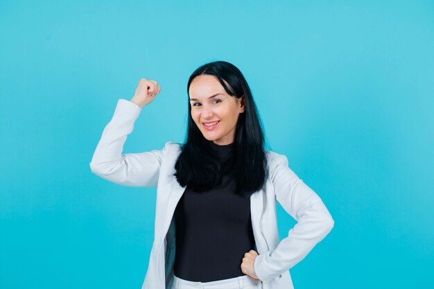 Smiling girl is raising up her fist and putting other hand on waist on blue background