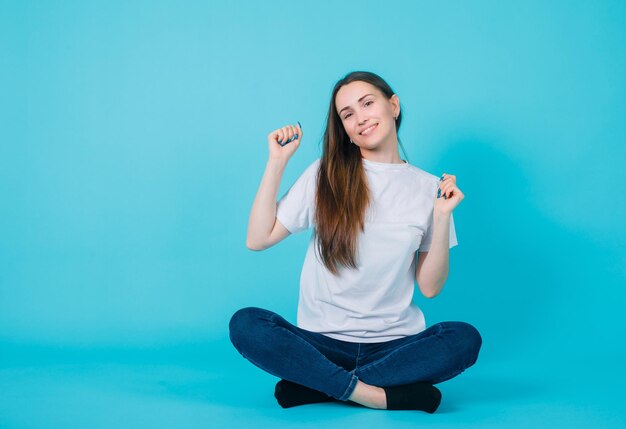 Smiling girl is raising up her fists by sitting on floor on blue background