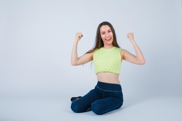 Smiling girl is raising up her fists by sitting on floor on white background