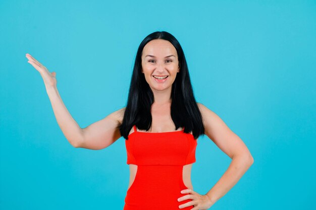 Smiling girl is raising up her hand and putting other hand on waist on blue background