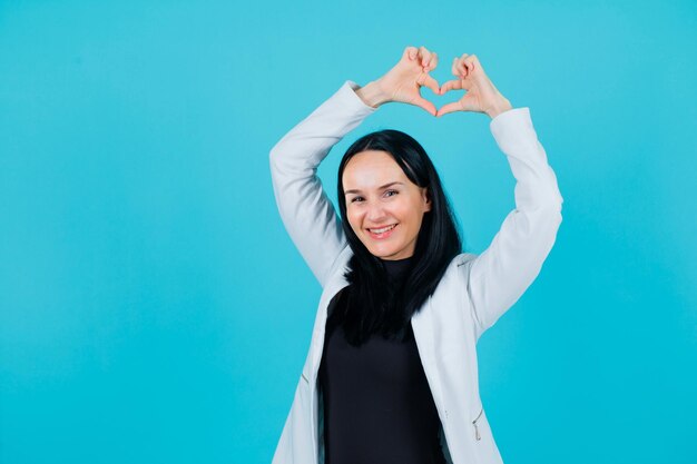 Smiling girl is showing heart gesture by raising up her hands above head on blue background