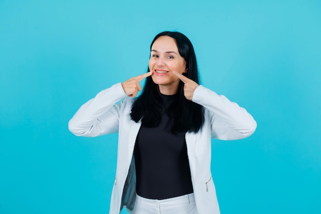 La ragazza sorridente sta mostrando il suo sorriso con gli indici su sfondo blu