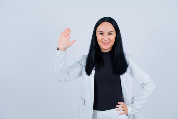 Smiling girl is showing hi gesture and putting other hand on waist on white background