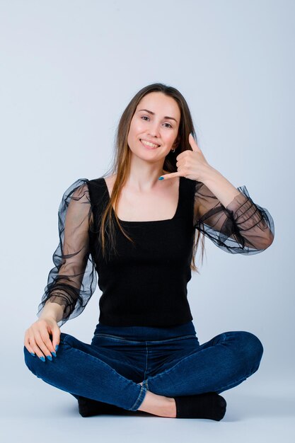 Smiling girl is showing phone gesture with hand by sitting on floor on white background