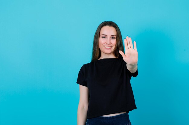 Smiling girl is showing stop gesture by extending hand to camera on blue background