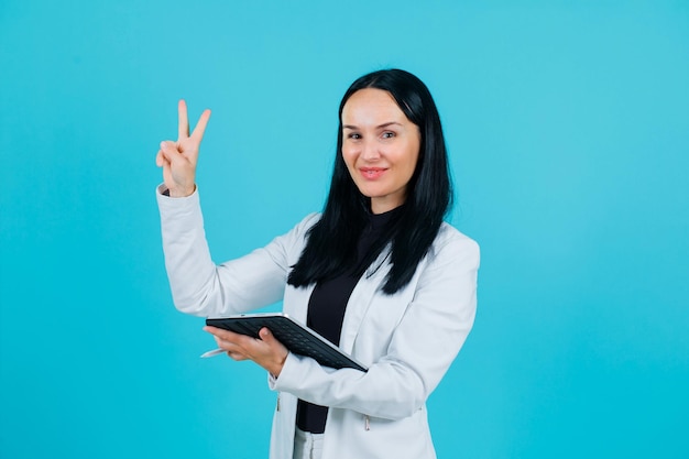 Smiling girl is showing two gesture by holding tablet on blue background