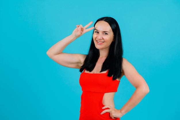 Smiling girl is showing two gesture and putting other hand on waist on blue background