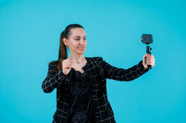 Smiling girl is taking selfie with her mini camera by raising up her fist on blue background
