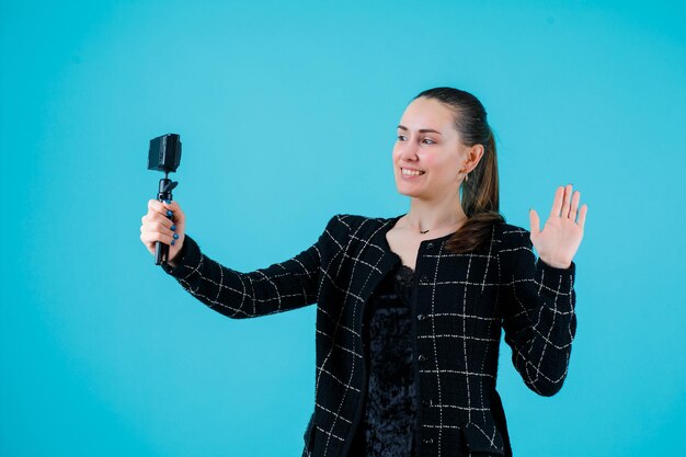 Smiling girl is taking selfie with her mini camera by showing hi gesture on blue background