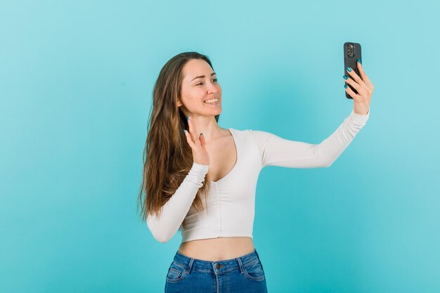 Smiling girl is talking on videocall by showing hi gesture to camera on blue background