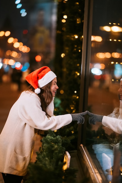 Smiling girl looking through shop window, city lights on background, in street of european city, wearing santa hat.