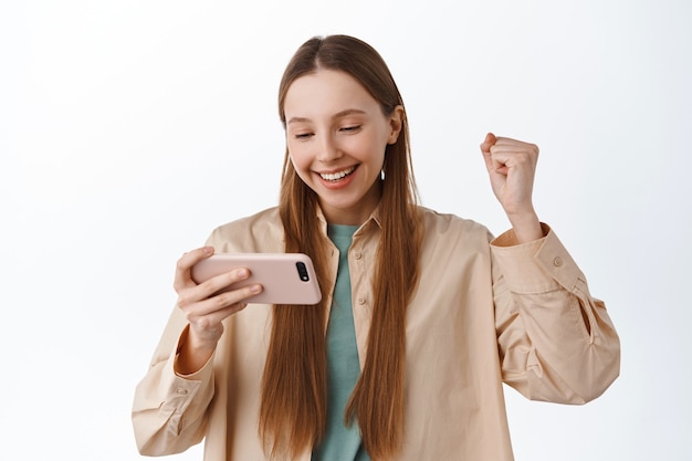 笑顔の女の子がスマートフォンを見て喜ぶ、ガッツポーズを喜ぶ、モバイルビデオゲームで勝つ、オンラインでの勝利を祝う、アプリで目標を達成する、白い壁の上に立つ