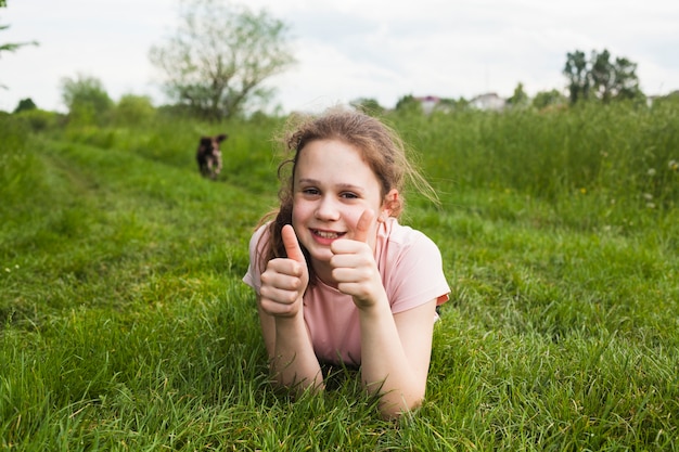Smiling girl lying on green grass and showing thumb up gesture in park