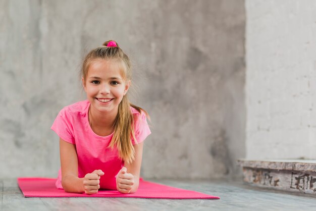 Smiling girl lying on pink exercise mat against concrete backdrop