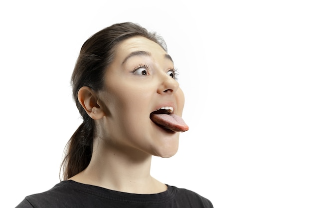 Smiling girl opening her mouth and showing the long big giant tongue isolated on white wall. Looks shocked, attracted, wondered and astonished. Copyspace for ad. Human emotions, marketing.