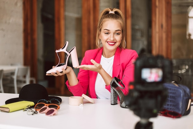Smiling girl in pink jacket happily showing shoes on heel while recording new fashion video for vlog on camera