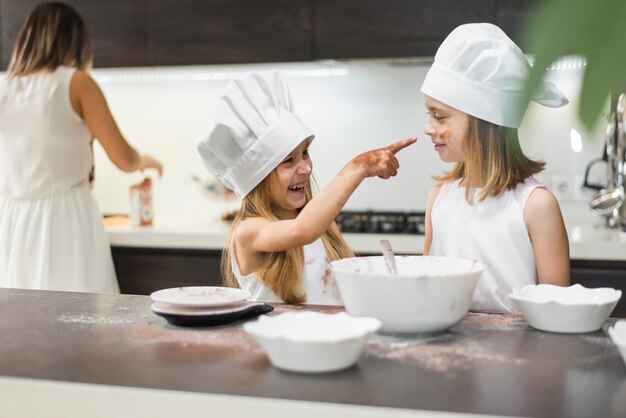 Smiling girl pointing her sister with messy hands in kitchen