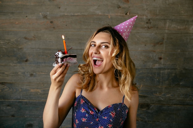 Ragazza sorridente pronta da mangiare un pezzo di torta di crema al cioccolato con candela, indossando il cappello festivo.