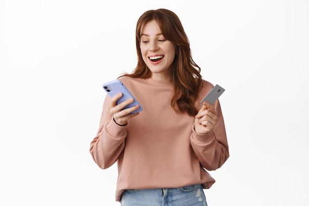 Smiling girl shops online, holding credit card and mobile phone, looking at smartphone screen with happy face, order something in internet, standing on white