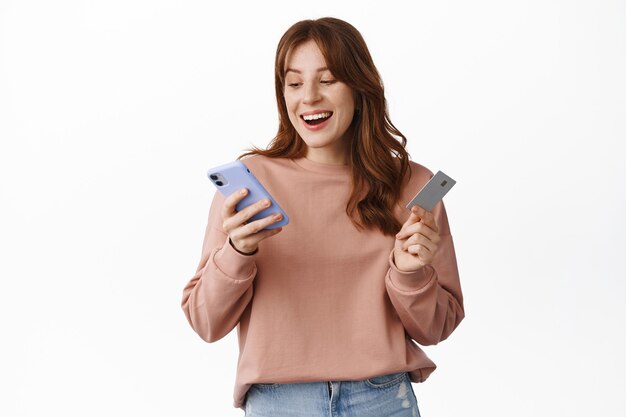 Smiling girl shops online, holding credit card and mobile phone, looking at smartphone screen with happy face, order something in internet, standing on white