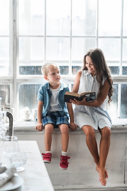 Foto gratuita ragazza sorridente che mostra libro al suo fratello piccolo che si siede vicino alla finestra