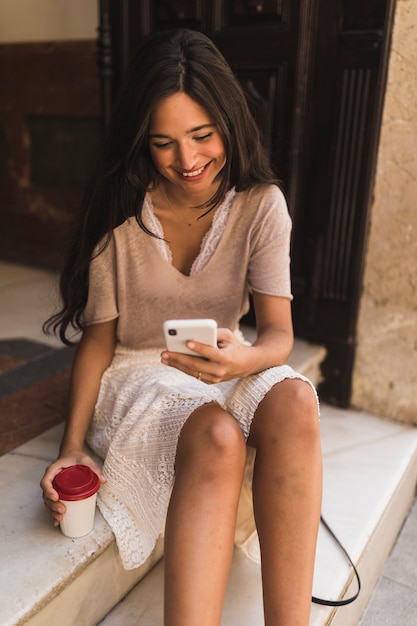 Free photo smiling girl sitting on steps holding disposable coffee cup using cell phone