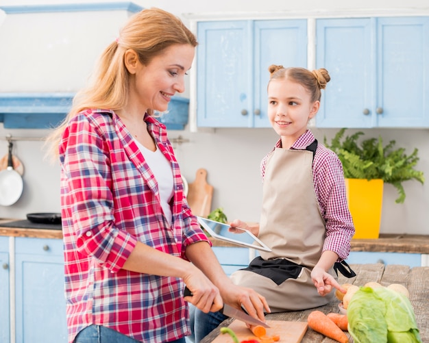 Smiling girl sitting on table looking at her mother cutting carrot with knife