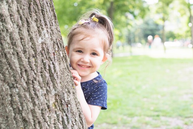 Smiling girl standing behind the tree trunk peeking in the garden